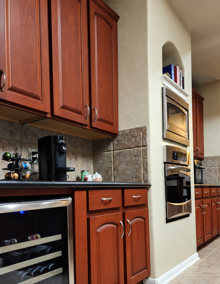 Reddish-brown kitchen cabinets around built-in ovens, a wine refrigerator, and a tiled backsplash.