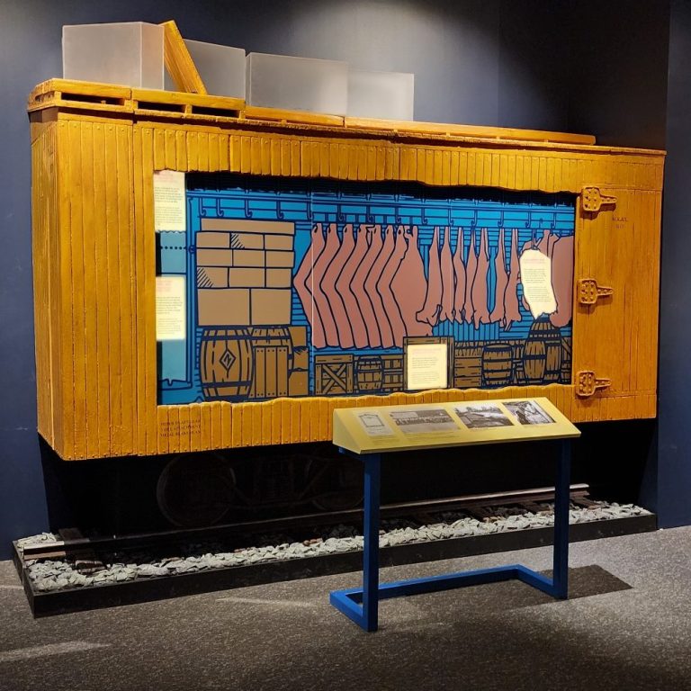 Half-scale wooden railcar exhibit with a cutaway side, display track, and interpretive panel.